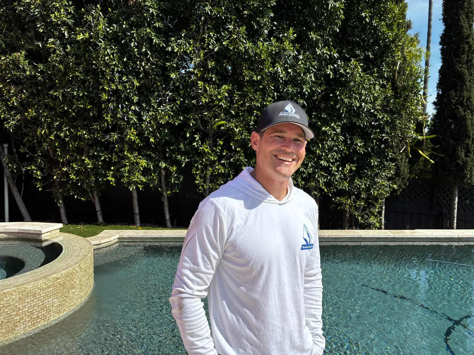 Brett Murdock smiling in a branded Murdock Pool Service hoodie beside an upscale customer pool and spa
